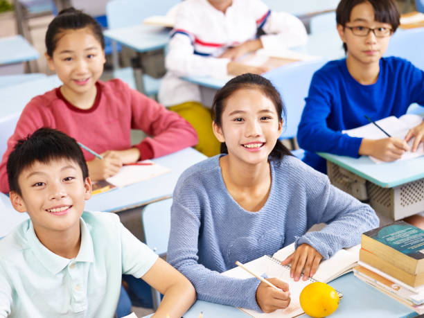 Four smiling students sit at desks in a classroom with books and notebooks.