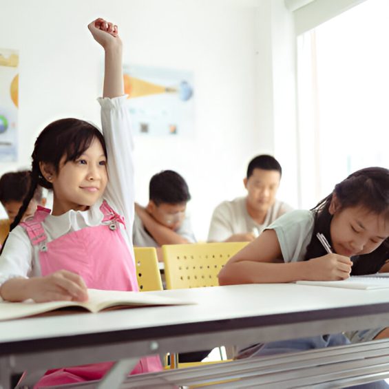 Young girl in a classroom raises her hand while other students write in notebooks.
